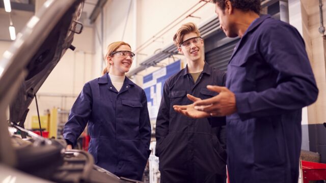 Male Tutor With Students Looking At Car Engine On Auto Mechanic Apprenticeship Course At College.