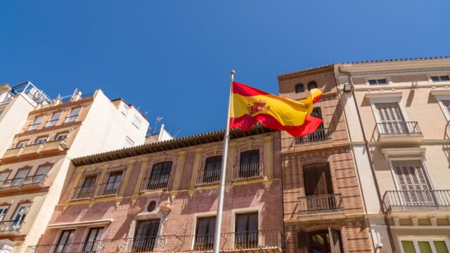 Spanish flag in the old town of the city of Malaga. Beautiful typically Spanish buildings architecture in background. Blue sky in a sunny summer day.