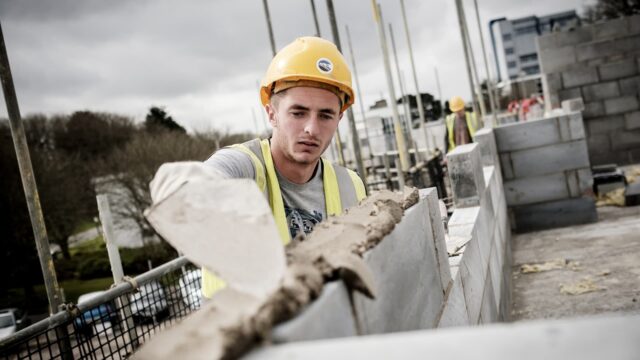 A bricklayer wearing a hard hat and high vis jacket is putting cement on bricks with a trowel.