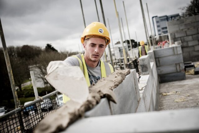 A bricklayer wearing a hard hat and high vis jacket is putting cement on bricks with a trowel.