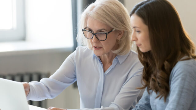 Two women look at a laptop together. They are both dressed formally.