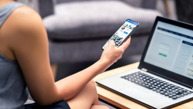 An unknown person looks at social media feed on their mobile phone with their laptop open in front of them.
