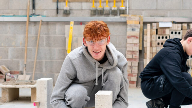 A bricklaying student crouches on the ground and looks towards the camera.