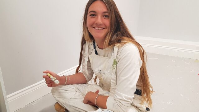 A student in overalls sits crossed legged on the floor with a paintbrush in hand.
