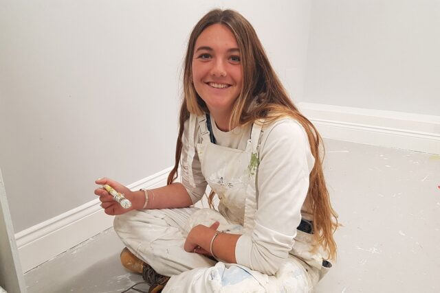 A student in overalls sits crossed legged on the floor with a paintbrush in hand.