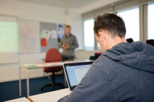 A student sits at a classroom table with a laptop in front of him.
