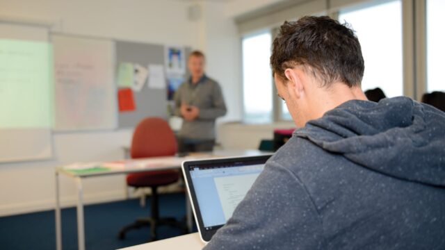A student sits at a classroom table with a laptop in front of him.
