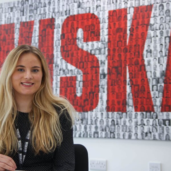 Chloe is sat at a desk with a large picture behind her that says 'WISKA'.