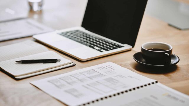A laptop, notebook, pen and coffee layed out on a table.