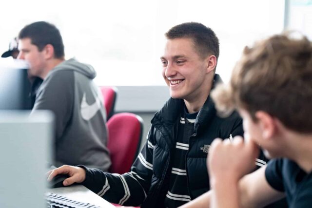 A student sat at a computer laughing with other students.