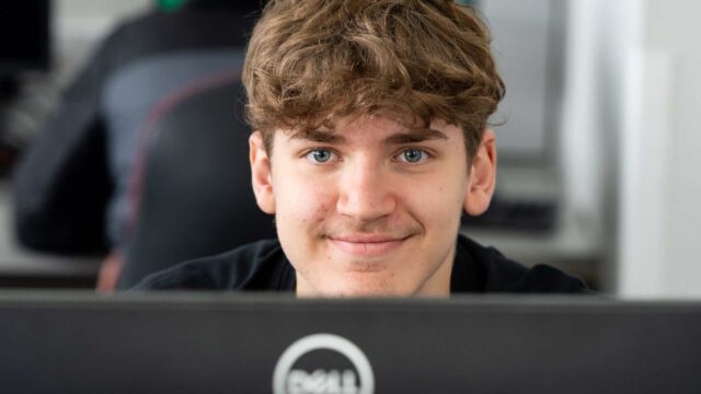A student looks over the top of his computer towards the camera and smiles.