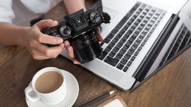 Photographer editing photos on laptop. There is a camera, coffe cup and pencil on the desk.
