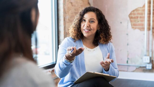 A woman sits opposite an unrecognisable person. She has a clipboard and pen.