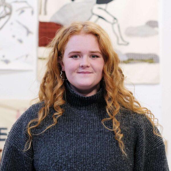 Ella has curly red hair and is wearing a grey roll neck jumper.