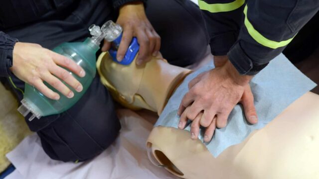 A person gives CPR to a dummy on the ground while another person holds a breathing apparatus to the dummy.