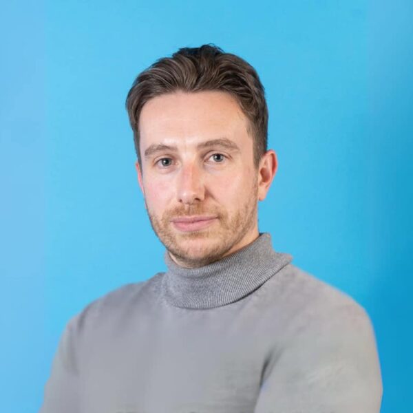 Nick is sat against a blue background and is wearing a grey jumper.