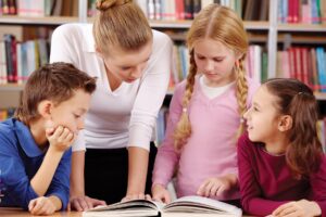 An adult stands with three children who are looking at a book together.