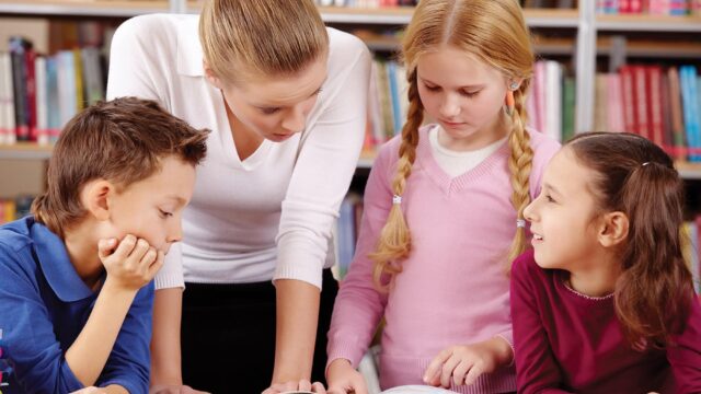 An adult stands with three children who are looking at a book together.