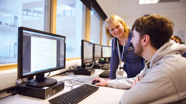 A student sits at a computer with a member of staff stood with them.