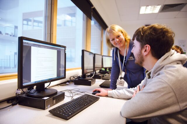 A student sits at a computer with a member of staff stood with them.