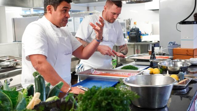 Paul Ainsworth giving a cooking demonstration. There are bowls and trays and food in the professional kitchen.