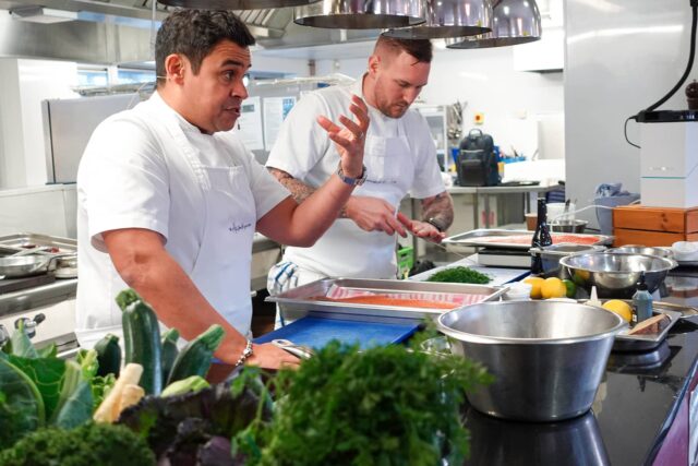 Paul Ainsworth giving a cooking demonstration. There are bowls and trays and food in the professional kitchen.