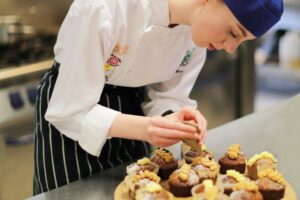 A student piping onto the top of small cakes in a kitchen.