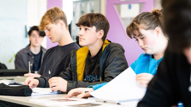 Students sit at classroom tables and face the front of the class.