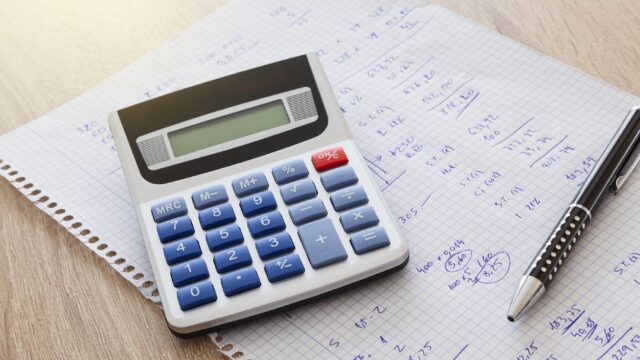 On a wooden table is a sheet of notebook squares with handwritten numbers and calculations and on the sheet is a pen and a calculator.