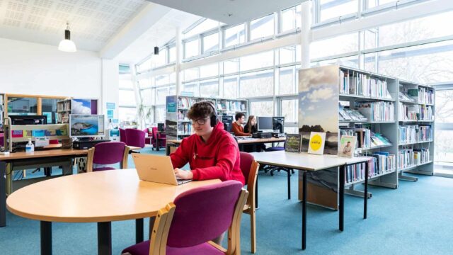 A library within the College. There are tables, chairs, bookcases and students sat at computers.