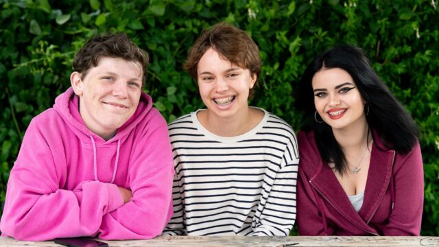Three students sit at a picnic bench facing the camera. All three of them smile.