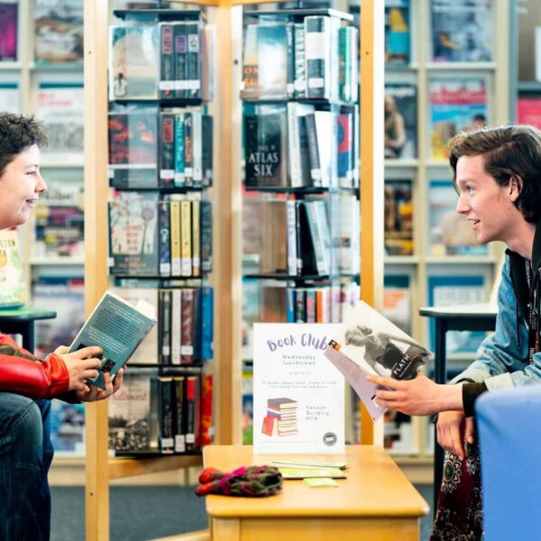 Two students sat in the College library with books open. A coffee table in between them with a poster that says Book Club.