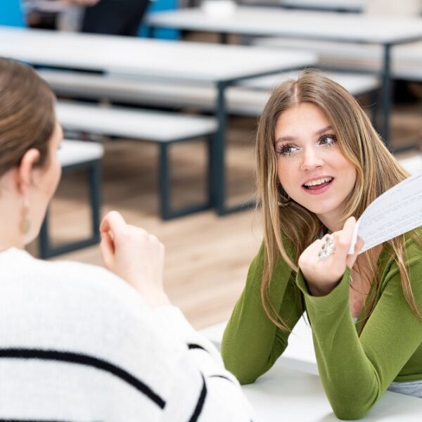 Two smiling students chat together at a table. One student holds a piece of paper in her hand.