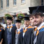 A group of graduates stand outside in Truro city in their caps and gowns and pose for the camera. They have their arms around each other. The colours of the robes are black, blue and orange.