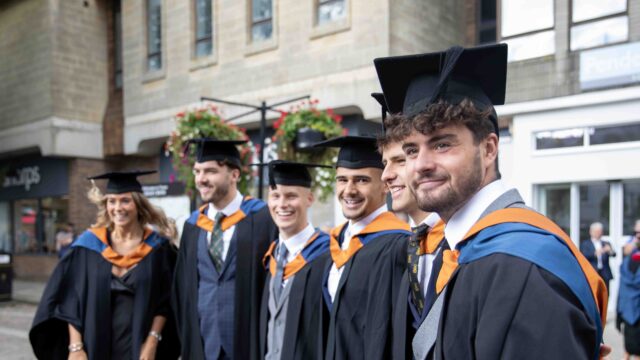 A group of graduates stand outside in Truro city in their caps and gowns and pose for the camera. They have their arms around each other. The colours of the robes are black, blue and orange.
