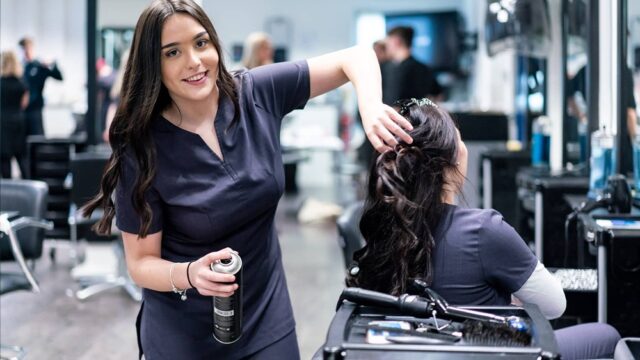 A hair and beauty student uses hairspray on another student within a professional salon environment.