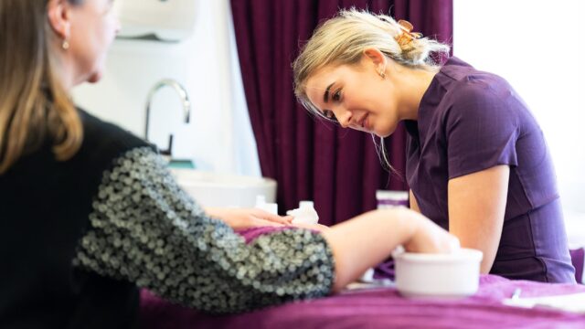 A student doing a nail treatment on a client. The student is wearing a purple tunic and there are purple towels on the work bench.