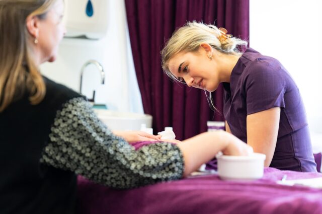 A student doing a nail treatment on a client. The student is wearing a purple tunic and there are purple towels on the work bench.