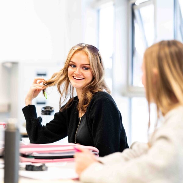 A student is playing with her hair as she is smiling at another student. They have pens and notepads in front of them.