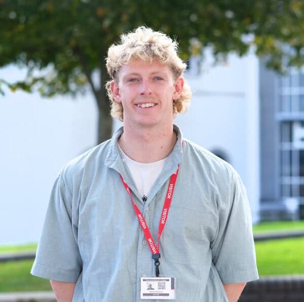 Henry is stood outside at the college with his hands behind his back and wearing a visitor lanyard.