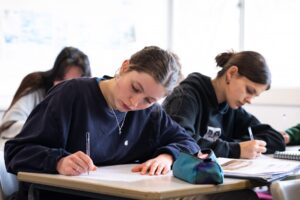 Students sit at classroom tables writing with pen on paper.