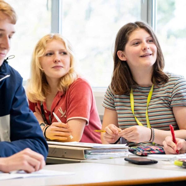 Students sat in a classroom look forwards to the front of the class. There are pencil cases, folders and pens on the tables.