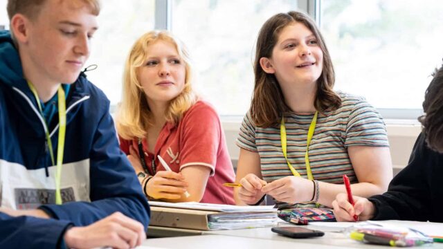 Students sat in a classroom look forwards to the front of the class. There are pencil cases, folders and pens on the tables.