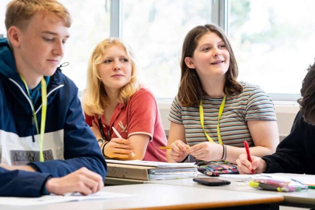 Students sat in a classroom look forwards to the front of the class. There are pencil cases, folders and pens on the tables.