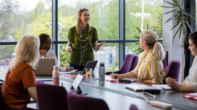 A wide-angle view of a group of a woman getting together for a business meeting in their place of work. The woman in the green blouse is leading the meeting and is encouraging the others to take part.