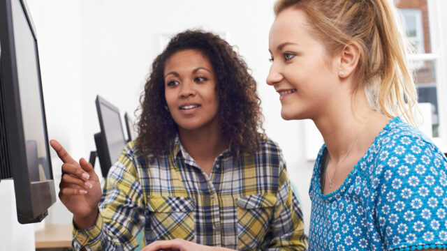 Two women look at a computer screen together. One woman points at the screen while the other is using a mouse.
