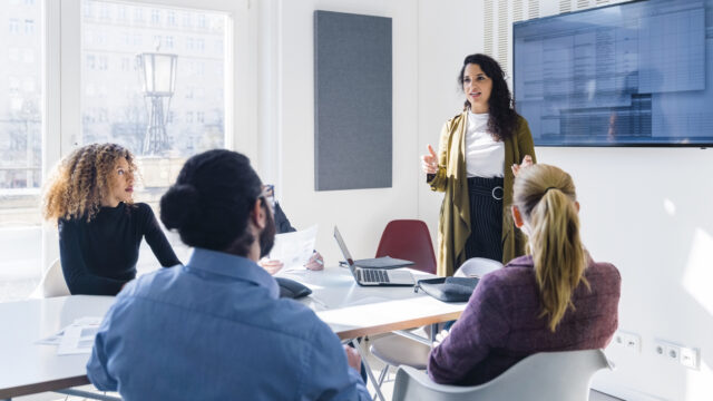 A woman stands in front of a small team in a brightly lit office. She is giving a presentation.