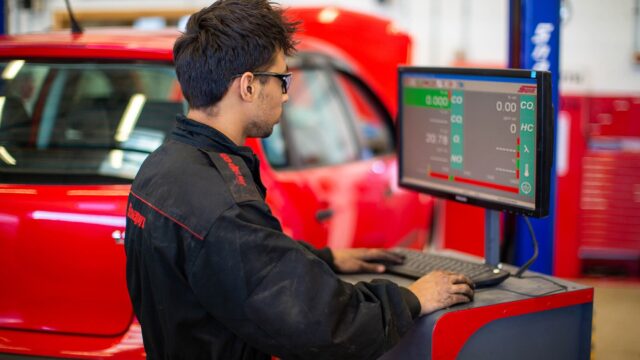 A student in overalls stands at a computer within a garage with a car in the background.