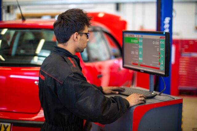 A student in overalls stands at a computer within a garage with a car in the background.