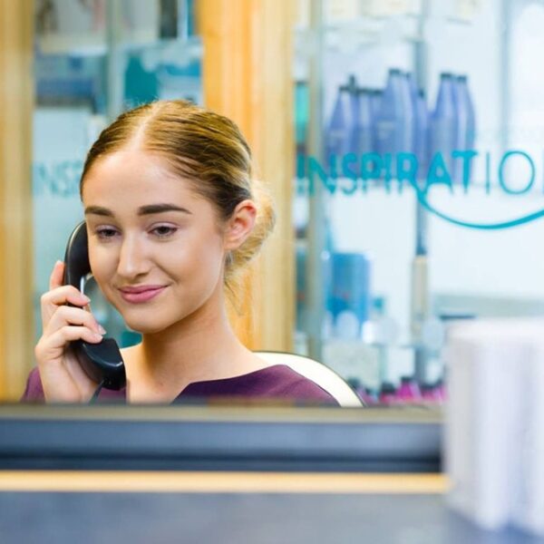A girl sits in the reception area of the inspiration salon and holds a phone to her ear. Her hair is tied back in a bun and she is wearing a purple tunic.